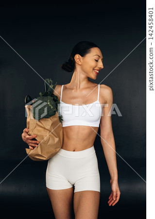 Female model in white sports bra and shorts holds a paper bag filled with fresh vegetables, showcasing a healthy lifestyle against a dark background Female model in white sports bra and shorts holds a paper bag filled with fresh vegetables, showcasing a healthy lifestyle against a dark background 142458401