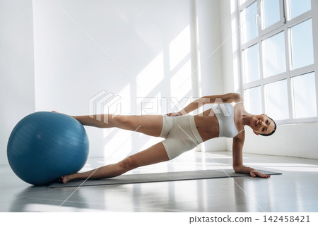 Slim female fitness model performing an exercise on a yoga mat with a blue stability ball in a bright, modern studio with large windows and natural light Slim female fitness model performing an exercise on a yoga mat with a blue stability ball in a bright, modern studio with large windows and natural light 142458421