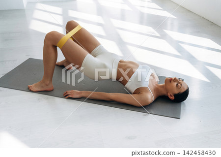 Slim female fitness model performing a bridge exercise on a yoga mat, wearing white athletic wear with a resistance band around her thighs in a bright indoor space Slim female fitness model performing a bridge exercise on a yoga mat, wearing white athletic wear with a resistance band around her thighs in a bright indoor space 142458430
