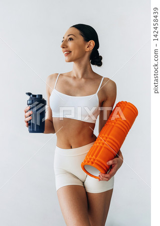 Slim female athlete in white sports bra and beige shorts holds a blue shaker bottle and an orange foam roller, posing against a plain white background Slim female athlete in white sports bra and beige shorts holds a blue shaker bottle and an orange foam roller, posing against a plain white background 142458439