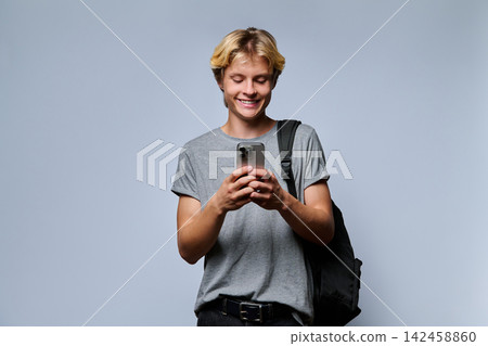 Smiling male student looking at smartphone with backpack on grey background Smiling male student looking at smartphone with backpack on grey background 142458860