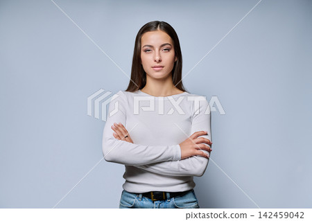 Serious young woman standing with crossed arms looking at camera on grey background Serious young woman standing with crossed arms looking at camera on grey background 142459042