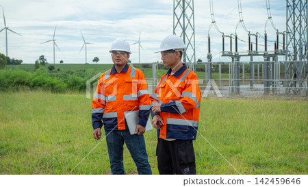 Two Asian male electrical engineers wearing hardhats and safety vests discuss work while holding laptop at power substation with wind turbines. Two Asian male electrical engineers wearing hardhats and safety vests discuss work while holding laptop at power substation with wind turbines. 142459646