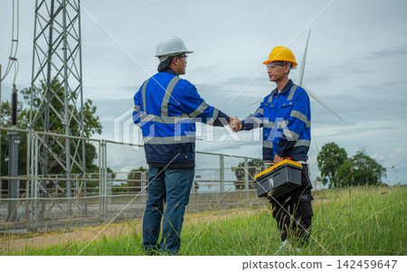 Two Asian male electrical engineers shaking hands at a wind turbine power station. Electrical technicians in hardhats partnership concept for clean energy. Two Asian male electrical engineers shaking hands at a wind turbine power station. Electrical technicians in hardhats partnership concept for clean energy. 142459647