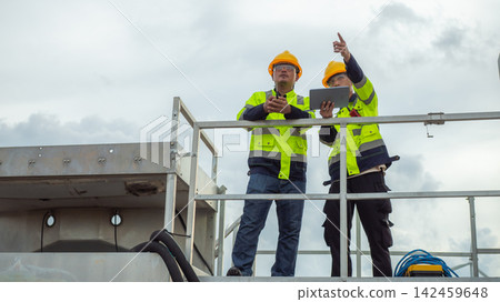 Two Asian male electrical engineers wearing hardhats and safety vests use tablet and walkie-talkie while pointing ahead on a high platform structure. Two Asian male electrical engineers wearing hardhats and safety vests use tablet and walkie-talkie while pointing ahead on a high platform structure. 142459648