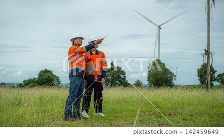 Two Asian male engineers holding a laptop point towards wind turbines in a green field. Renewable energy technicians inspect electric power station. Two Asian male engineers holding a laptop point towards wind turbines in a green field. Renewable energy technicians inspect electric power station. 142459649