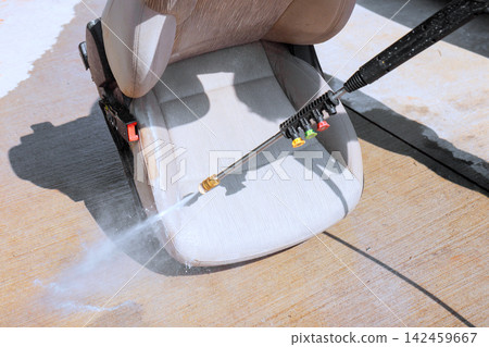 Man uses pressure washer to clean car seat in driveway water sprays onto fabric. Man uses pressure washer to clean car seat in driveway water sprays onto fabric. 142459667