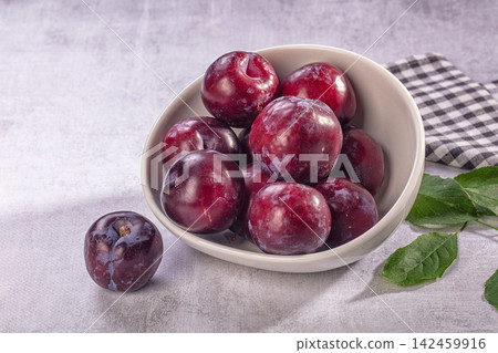 Fresh plums in ceramic bowl with green leaves and checkered cloth on gray surface Fresh plums in ceramic bowl with green leaves and checkered cloth on gray surface 142459916