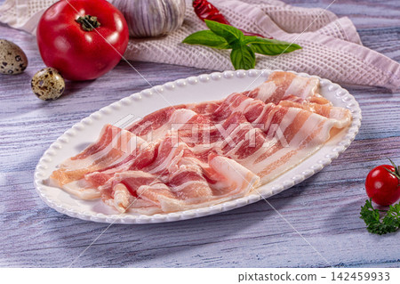 Raw bacon strips on white plate with tomato garlic and herbs on rustic wood Raw bacon strips on white plate with tomato garlic and herbs on rustic wood 142459933