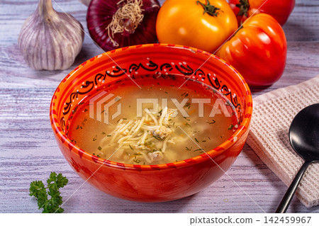 Chicken noodle soup in ornate red bowl with fresh vegetables on rustic wooden table Chicken noodle soup in ornate red bowl with fresh vegetables on rustic wooden table 142459967