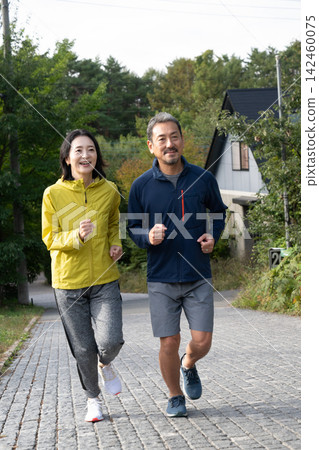 A middle-aged couple running or jogging side-by-side along a path dappled with sunlight filtering through the trees. A middle-aged couple running or jogging side-by-side along a path dappled with sunlight filtering through the trees. 142460075