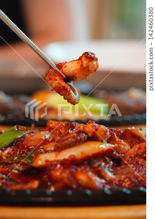 Chopsticks picking up spicy stir-fried octopus with vegetables from a sizzling hot skillet, fiery red Korean street food style close-up Chopsticks picking up spicy stir-fried octopus with vegetables from a sizzling hot skillet, fiery red Korean street food style close-up 142460380