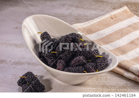 Fresh dark mulberries in white ceramic bowl on textured surface with striped cloth Fresh dark mulberries in white ceramic bowl on textured surface with striped cloth 142461184