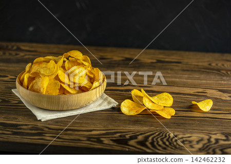 Potato chips in bowl on a wooden background. Salty crisps scattered on a table Potato chips in bowl on a wooden background. Salty crisps scattered on a table 142462232