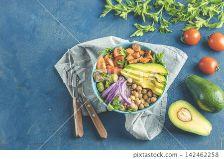 Healthy food. Healthy vegan lunch bowl of fried sweet potato, chickpeas, red onion, avocado, tomatoes, and brussels sprouts. Top view, classic blue concrete surface, flat lay Healthy food. Healthy vegan lunch bowl of fried sweet potato, chickpeas, red onion, avocado, tomatoes, and brussels sprouts. Top view, classic blue concrete surface, flat lay 142462258