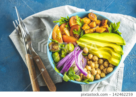 Healthy food. Healthy vegan lunch bowl of fried sweet potato, chickpeas, red onion, avocado, tomatoes, and brussels sprouts. Top view, classic blue concrete surface, flat lay Healthy food. Healthy vegan lunch bowl of fried sweet potato, chickpeas, red onion, avocado, tomatoes, and brussels sprouts. Top view, classic blue concrete surface, flat lay 142462259