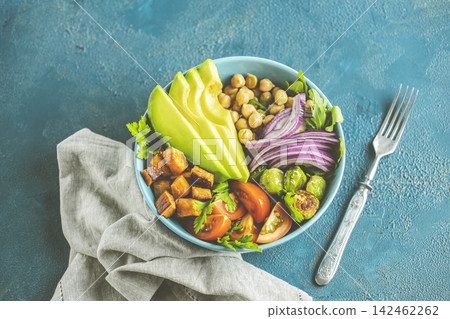 Healthy food. Healthy vegan lunch bowl of fried sweet potato, chickpeas, red onion, avocado, tomatoes, and brussels sprouts. Top view, classic blue concrete surface, flat lay Healthy food. Healthy vegan lunch bowl of fried sweet potato, chickpeas, red onion, avocado, tomatoes, and brussels sprouts. Top view, classic blue concrete surface, flat lay 142462262
