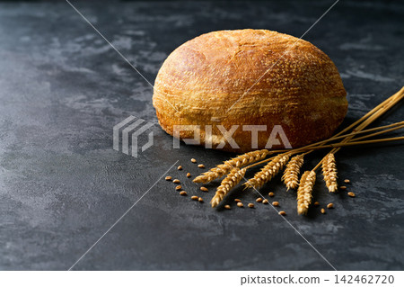 Round loaf of wheat rustic bread with sourdough and ears of wheat on dark kitchen table, selective focus. Round loaf of wheat rustic bread with sourdough and ears of wheat on dark kitchen table, selective focus. 142462720