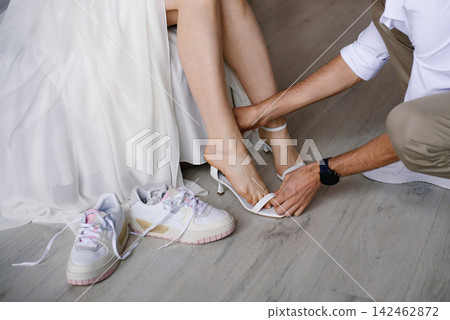 Groom Helping Bride to Put on White High Heel Shoes with Sneakers on Floor Groom Helping Bride to Put on White High Heel Shoes with Sneakers on Floor 142462872