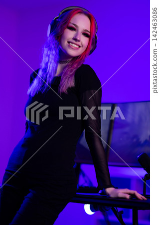 Smiling young woman streaming with headphones and gaming setup in purple lighting Smiling young woman streaming with headphones and gaming setup in purple lighting 142463086