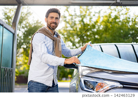 A smiling man in casual attire is carefully drying the hood of his car with a bright blue microfiber towel after a wash A smiling man in casual attire is carefully drying the hood of his car with a bright blue microfiber towel after a wash 142463129