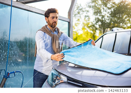A caucasian man is carefully drying his car with a blue microfiber cloth after washing it at an outdoor car wash station A caucasian man is carefully drying his car with a blue microfiber cloth after washing it at an outdoor car wash station 142463133