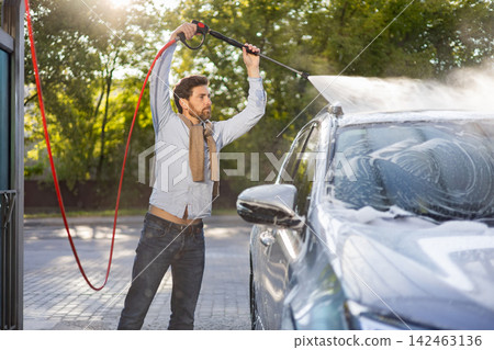 A man is actively cleaning his vehicle using a high-pressure water sprayer, creating a foamy lather on the car's surface A man is actively cleaning his vehicle using a high-pressure water sprayer, creating a foamy lather on the car's surface 142463136