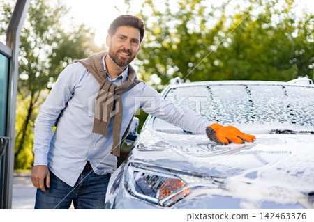 A smiling caucasian man wearing an orange glove washes his car with soap and water outdoors on a sunny day A smiling caucasian man wearing an orange glove washes his car with soap and water outdoors on a sunny day 142463376