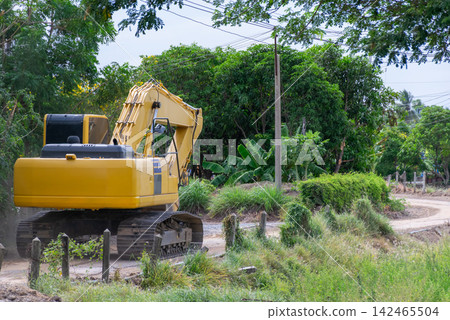 Large yellow excavator parked rural dirt road surrounded by lush tropical vegetation trees utility lines showing heavy machinery used for earthwork construction Large yellow excavator parked rural dirt road surrounded by lush tropical vegetation trees utility lines showing heavy machinery used for earthwork construction 142465504