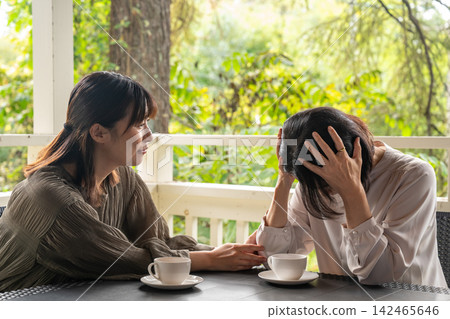 A woman comforts her depressed friend in a cafe surrounded by trees. A woman comforts her depressed friend in a cafe surrounded by trees. 142465646