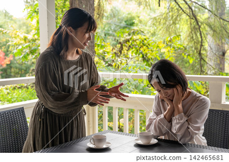 A woman confides her troubles to her friend in a cafe surrounded by trees, while her friend comforts her. A woman confides her troubles to her friend in a cafe surrounded by trees, while her friend comforts her. 142465681