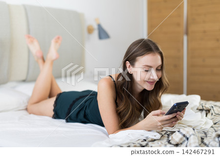 Adult woman scrolling smartphone sitting on bed Adult woman scrolling smartphone sitting on bed 142467291