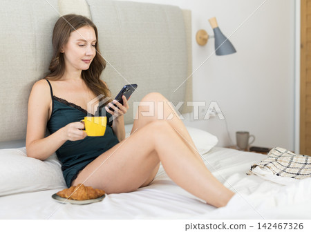 Woman in bedroom with cup of coffee and croissant, holding phone Woman in bedroom with cup of coffee and croissant, holding phone 142467326
