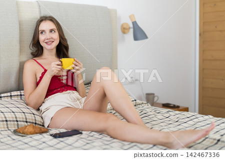 Adult woman drinking coffee sitting on bed in bedroom Adult woman drinking coffee sitting on bed in bedroom 142467336