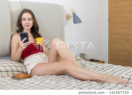 Woman drinking morning coffee and looking at smartphone screen in bedroom 142467339
