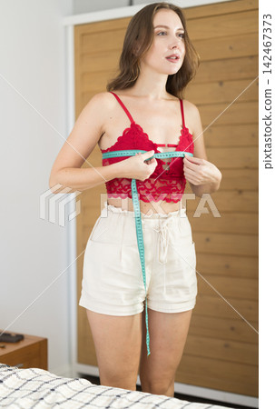 woman stands in the bedroom and thoughtfully measures her figure with a measuring tape 142467373