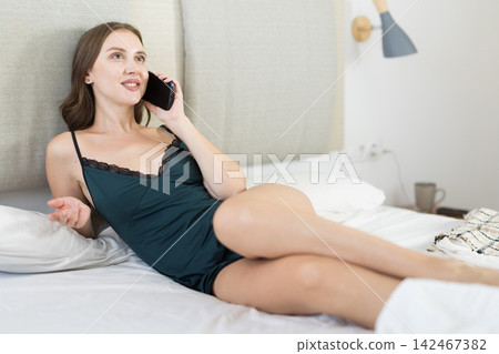 Adult woman talking on smartphone sitting on bed Adult woman talking on smartphone sitting on bed 142467382