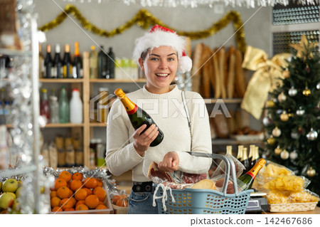 Feale customer in Santa Claus hat with cart of groceries buys bottle of champagne in supermarket - prepares to celebrate Christmas Feale customer in Santa Claus hat with cart of groceries buys bottle of champagne in supermarket - prepares to celebrate Christmas 142467686