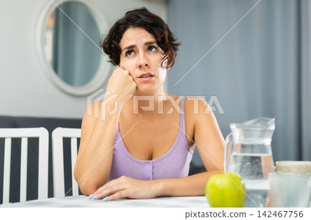 Dissatisfied young woman sitting at the table in room of her apartment Dissatisfied young woman sitting at the table in room of her apartment 142467756
