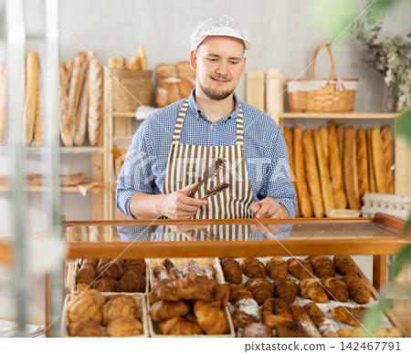 Positive bakery seller with tongs in his hands offers to buy fresh croissants Positive bakery seller with tongs in his hands offers to buy fresh croissants 142467791