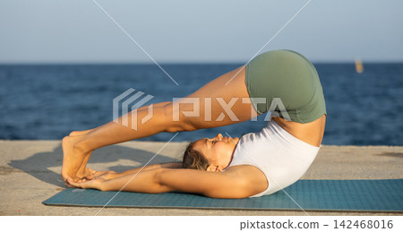 On windy day young slender woman does yoga near sea doing halasana On windy day young slender woman does yoga near sea doing halasana 142468016