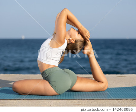 Flexible fit woman doing yoga on the seashore, performing seated back-bending asana Rajakapotasana, or King Pigeon Pose Flexible fit woman doing yoga on the seashore, performing seated back-bending asana Rajakapotasana, or King Pigeon Pose 142468096