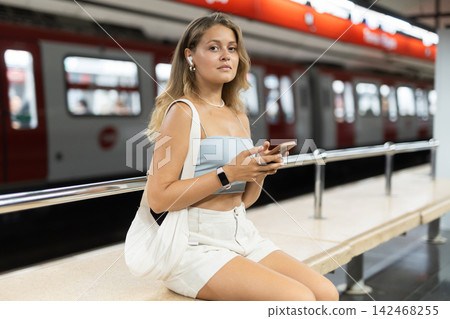 Young girl sitting in subway platform holding phone and wireless headphones on airs Young girl sitting in subway platform holding phone and wireless headphones on airs 142468255