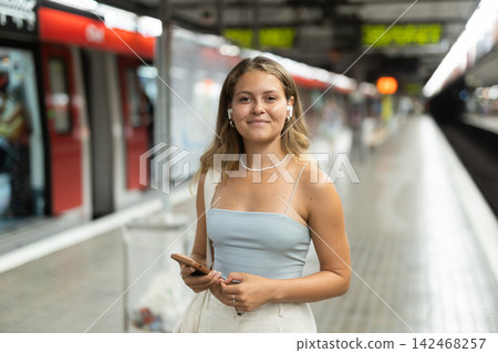 Positive young girl standing in platform holding phone with wireless headphones on airs Positive young girl standing in platform holding phone with wireless headphones on airs 142468257