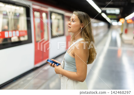Girl with phone is standing on subway platform, waiting for train to arrive Girl with phone is standing on subway platform, waiting for train to arrive 142468297