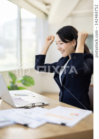 A young Japanese businesswoman makes a triumphant gesture while looking at her computer at her desk by the window. A young Japanese businesswoman makes a triumphant gesture while looking at her computer at her desk by the window. 142468752
