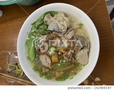 A white bowl of clear broth noodle soup with sliced meat, leafy greens, chopped scallions, and crispy garnish sits on a wooden table beside chopsticks. A white bowl of clear broth noodle soup with sliced meat, leafy greens, chopped scallions, and crispy garnish sits on a wooden table beside chopsticks. 142469147