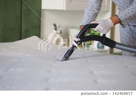 A woman is using a vacuum cleaner on a mattress in a bedroom. A woman is using a vacuum cleaner on a mattress in a bedroom. 142469657