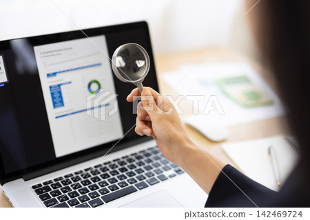 特寫鏡頭拍攝了一位年輕的日本女商人，她正拿著放大鏡對著筆記型電腦螢幕。 142469724