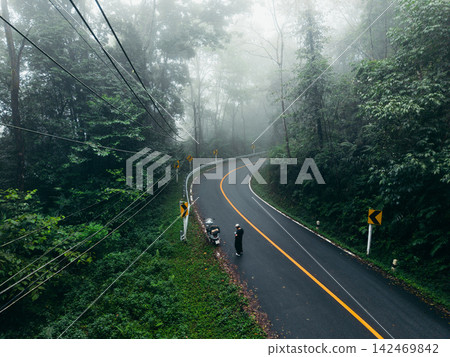 Aerial view of a traveler with a motorcycle stopped on a winding misty forest road. 142469842
